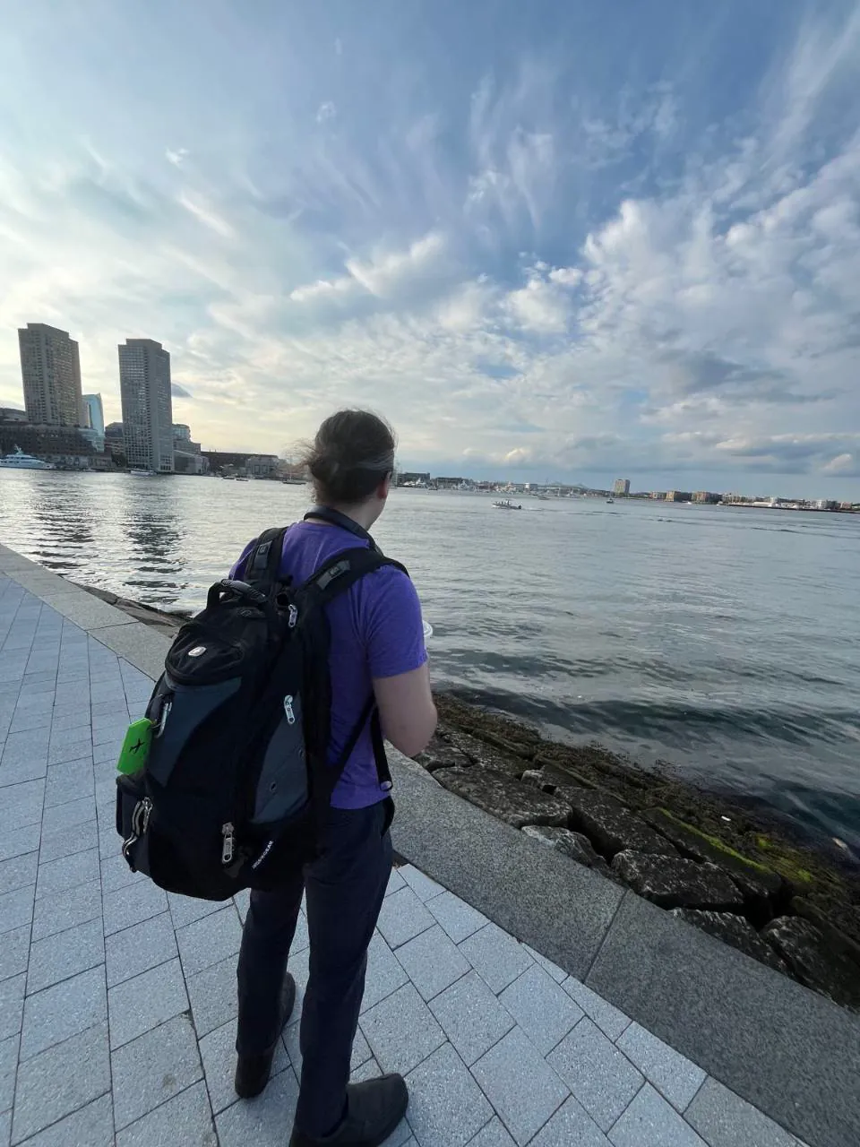 Andrew standing beside a waterfront, viewed from behind beneath a wide cloudy sky.